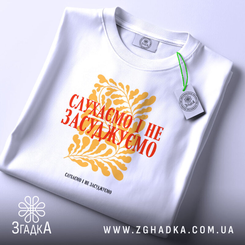 Футболка слухаємо і не засуджуємо, біла, складена на поверхні, фон світло-сірий, принт жовтий з рослинним орнаментом та червоним текстом, на комірці є етикетка.