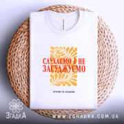 Футболка слухаємо і не засуджуємо, біла, складена на плетеній кошику, фон білий, принт жовтий з рослинним орнаментом та червоним текстом.