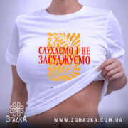 Футболка слухаємо і не засуджуємо, біла, складена на поверхні, фон світло-сірий, принт жовтий з рослинним орнаментом та червоним текстом, на комірці є етикетка.