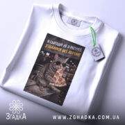 Футболка сьогодні не в ресурсі. Біла футболка, зображення кота в окулярах з шматком піци, розташоване на футболці, яка лежить на світлому фоні. На футболці є етикетка з зеленим шнурком.