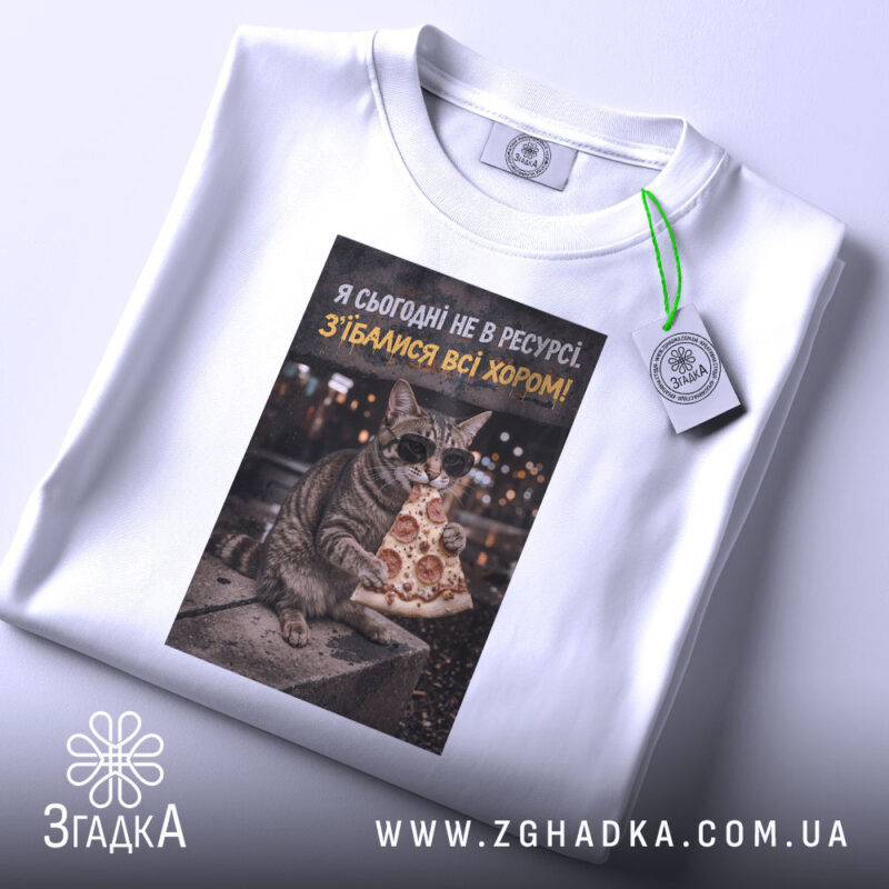 Футболка сьогодні не в ресурсі. Біла футболка, зображення кота в окулярах з шматком піци, розташоване на футболці, яка лежить на світлому фоні. На футболці є етикетка з зеленим шнурком.