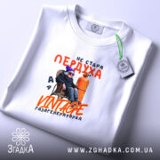 Футболка стара пердуха, біла, зображена на плоскій поверхні, фон світло-фіолетовий, принт з текстом "не стара пердуха" та зображенням персонажа в окулярах і шапці, що сидить на стільці біля газового балона, з етикеткою на комірці.