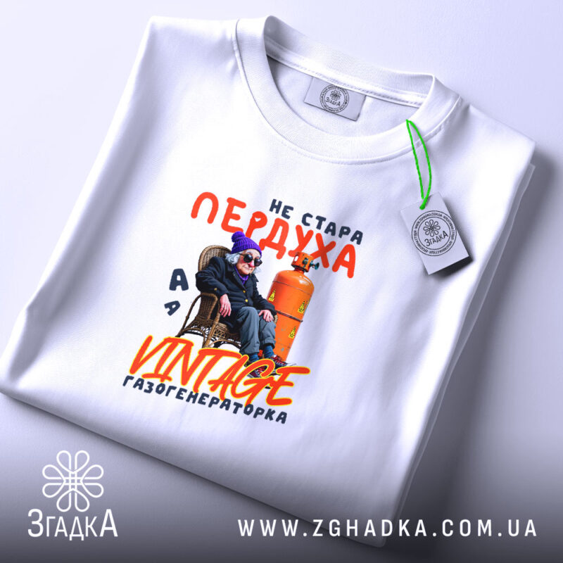 Футболка стара пердуха, біла, зображена на плоскій поверхні, фон світло-фіолетовий, принт з текстом "не стара пердуха" та зображенням персонажа в окулярах і шапці, що сидить на стільці біля газового балона, з етикеткою на комірці.