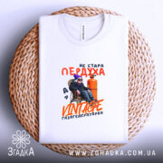 Футболка стара пердуха, біла, складена на плоскій поверхні, фон світло-фіолетовий, принт з текстом "не стара пердуха" та зображенням персонажа в окулярах і шапці, що сидить на стільці біля газового балона, розташована на плетеній основі.