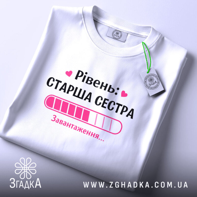 Футболка старша сестра, біла, на столі, з етикеткою, фон світло-сірий, принт з написом 'Рівень: СТАРША СЕСТРА' та графіком завантаження в рожевих відтінках.