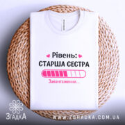 Футболка старша сестра, біла, на плетеній кошику, фон світло-сірий, принт з написом 'Рівень: СТАРША СЕСТРА' та графіком завантаження в рожевих відтінках.