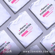 Футболка старша сестра, біла, на моделі, з легким макіяжем, фон світло-фіолетовий, принт з написом 'Рівень: СТАРША СЕСТРА' та графіком завантаження в рожевих відтінках.