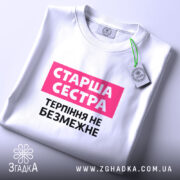 Футболка старша сестра, біла, на столі, з фоном у світло-сірому кольорі, принт з написом 'Старша сестра' у рожевому прямокутнику та 'Терпіння не безмежне' чорним шрифтом, з етикеткою на зеленому шнурку.