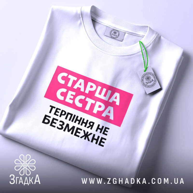 Футболка старша сестра, біла, на столі, з фоном у світло-сірому кольорі, принт з написом 'Старша сестра' у рожевому прямокутнику та 'Терпіння не безмежне' чорним шрифтом, з етикеткою на зеленому шнурку.