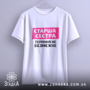 Футболка старша сестра, біла, підвішена на вішалці, з фоном у світло-сірому кольорі, принт з написом 'Старша сестра' у рожевому прямокутнику та 'Терпіння не безмежне' чорним шрифтом.