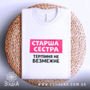 Футболка старша сестра, біла, на столі, з фоном у світло-сірому кольорі, складена на плетеній підставці, принт з написом 'Старша сестра' у рожевому прямокутнику та 'Терпіння не безмежне' чорним шрифтом.