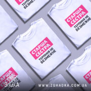 Футболка старша сестра, біла, на моделі, з фоном у світло-фіолетових тонах, принт з написом 'Старша сестра' у рожевому прямокутнику та 'Терпіння не безмежне' чорним шрифтом.