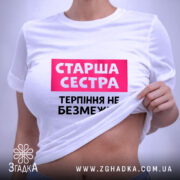 Футболка старша сестра, біла, на столі, з фоном у світло-сірому кольорі, принт з написом 'Старша сестра' у рожевому прямокутнику та 'Терпіння не безмежне' чорним шрифтом, з етикеткою на зеленому шнурку.