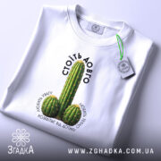 Футболка Стоїть довго, біла, на плоскій поверхні, з принтом кактусів, фон світло-сірий, етикетка з зеленим шнурком, текст над принтом чорний, напис 'СТОЇТЬ ДОВГО' розташований зверху.