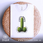 Футболка Стоїть довго, біла, на плоскій поверхні, з принтом кактусів, фон світлий, футболка розташована на плетеній основі, текст над принтом чорний, напис 'СТОЇТЬ ДОВГО' розташований зверху.