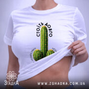 Футболка Стоїть довго, біла, на плоскій поверхні, з принтом кактусів, фон світло-сірий, етикетка з зеленим шнурком, текст над принтом чорний, напис 'СТОЇТЬ ДОВГО' розташований зверху.