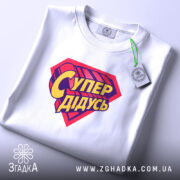 Футболка супер дідусь, біла, складена на поверхні, з верхнього ракурсу, фон світло-сірий, принт червоного кольору з жовтим написом 'Супер дідусь', на етикетці зелена стрічка.