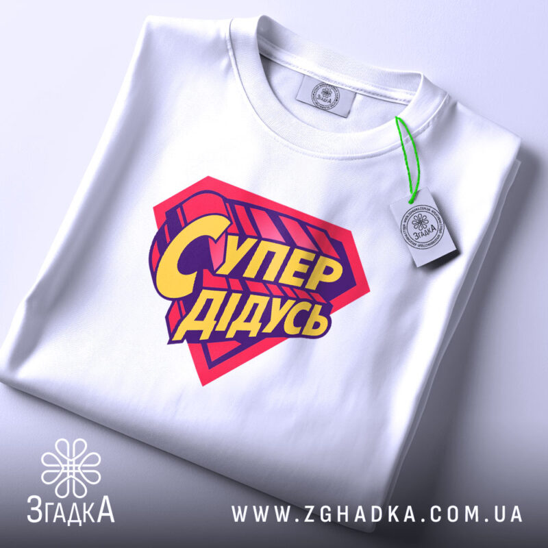 Футболка супер дідусь, біла, складена на поверхні, з верхнього ракурсу, фон світло-сірий, принт червоного кольору з жовтим написом 'Супер дідусь', на етикетці зелена стрічка.