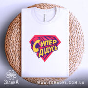 Футболка супер дідусь, біла, складена на плетеній кошику, з верхнього ракурсу, фон світло-сірий, принт червоного кольору з жовтим написом 'Супер дідусь'.