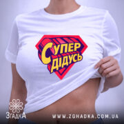 Футболка супер дідусь, біла, складена на поверхні, з верхнього ракурсу, фон світло-сірий, принт червоного кольору з жовтим написом 'Супер дідусь', на етикетці зелена стрічка.