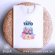 Футболка супер тато білого кольору, складена на плоскій поверхні з плетеного кошика, фон білий, принт зображує батька і сина у супергеройських костюмах з квітами.