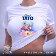 Футболка супер тато білого кольору, складена на плоскій поверхні, фон світло-фіолетовий, принт зображує батька і сина у супергеройських костюмах з квітами, на комірці є етикетка.