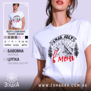 Футболка сила лісу в мені білого кольору, зображена на моделі, яка позує в три чверті, фон м'який пастельний, принт з горами та деревами в чорному кольорі з червоним написом.