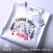 Футболка сила лісу в мені білого кольору, лежить на поверхні, ракурс зверху, фон світлий, принт з горами та деревами в чорному кольорі з червоним написом, на комірці є етикетка.