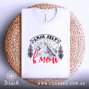 Футболка сила лісу в мені білого кольору, складена на плетеній основі, фон світлий, принт з горами та деревами в чорному кольорі з червоним написом, видно деталі комірця.