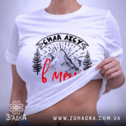 Футболка сила лісу в мені білого кольору, лежить на поверхні, ракурс зверху, фон світлий, принт з горами та деревами в чорному кольорі з червоним написом, на комірці є етикетка.