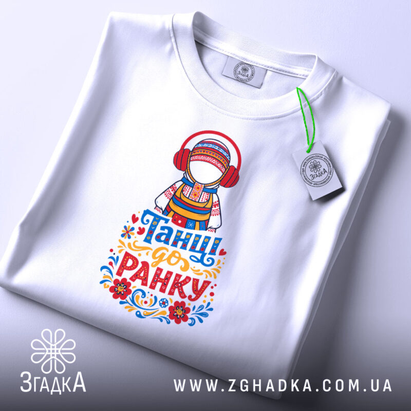 Футболка танці до ранку, біла, зображена зверху, фон світло-сірий, принт з дівчиною в національному вбранні з навушниками, яскраві кольори принта, текст 'Танці до ранку' внизу, на етикетці видно логотип.