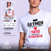 Футболка Тато білого кольору, зображена на молодому чоловікові, ракурс з близької відстані, фон світло-сірий, принт містить чорний напис "БЕТМЕН" та червоний напис "ТАТО справжній СУПЕРГЕРОЙ".