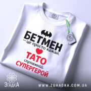 Футболка Тато білого кольору, зображена на столі, ракурс зверху, фон світло-сірий, принт містить чорний напис "БЕТМЕН" та червоний напис "ТАТО справжній СУПЕРГЕРОЙ", на футболці є етикетка.