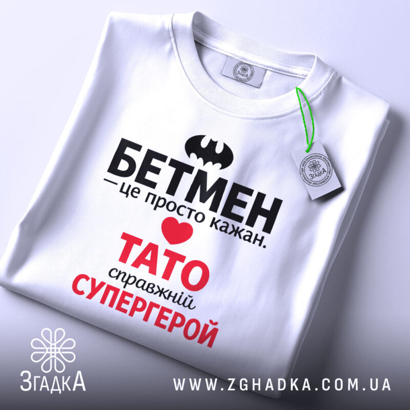 Футболка Тато білого кольору, зображена на столі, ракурс зверху, фон світло-сірий, принт містить чорний напис "БЕТМЕН" та червоний напис "ТАТО справжній СУПЕРГЕРОЙ", на футболці є етикетка.