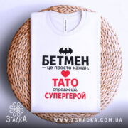 Футболка Тато білого кольору, зображена на плетеній основі, ракурс зверху, фон світло-сірий, принт містить чорний напис "БЕТМЕН" та червоний напис "ТАТО справжній СУПЕРГЕРОЙ".