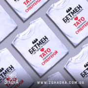 Футболка Тато білого кольору, зображена на молодому чоловікові, ракурс з близької відстані, фон світло-сірий, принт містить чорний напис "БЕТМЕН" та червоний напис "ТАТО справжній СУПЕРГЕРОЙ".