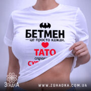 Футболка Тато білого кольору, зображена на столі, ракурс зверху, фон світло-сірий, принт містить чорний напис "БЕТМЕН" та червоний напис "ТАТО справжній СУПЕРГЕРОЙ", на футболці є етикетка.