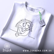 Футболка Терміново потрібні обійми, біла, складена на поверхні, фон світло-сірий, принт з контурним малюнком двох людей, що обіймаються, текст розташований у верхній частині, етикетка з зеленим шнурком.