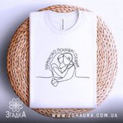 Футболка Терміново потрібні обійми, біла, складена на плетеній кошику, фон світлий, принт з контурним малюнком двох людей, що обіймаються, текст розташований у верхній частині.