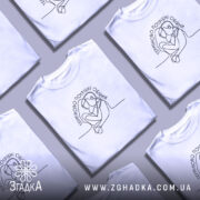 Футболка Терміново потрібні обійми, біла, зображена на моделі, яка позує з легким усмішкою, фон світло-фіолетовий, принт з контурним малюнком двох людей, що обіймаються, текст зверху написаний елегантним шрифтом.