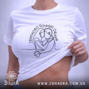 Футболка Терміново потрібні обійми, біла, складена на поверхні, фон світло-сірий, принт з контурним малюнком двох людей, що обіймаються, текст розташований у верхній частині, етикетка з зеленим шнурком.