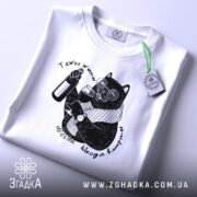 Футболка тяжко жити шкода вмерти, біла, складена на поверхні, ракурс зверху, фон світло-сірий, принт з чорним котом у окулярах, що тримає пляшку та келих, з етикеткою на рукаві.