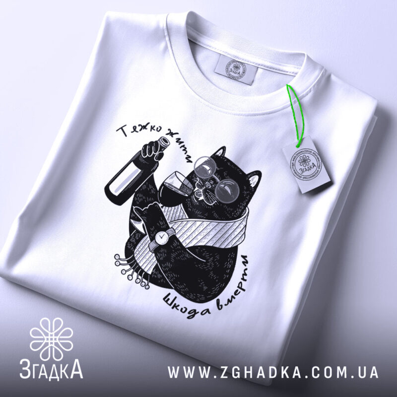 Футболка тяжко жити шкода вмерти, біла, складена на поверхні, ракурс зверху, фон світло-сірий, принт з чорним котом у окулярах, що тримає пляшку та келих, з етикеткою на рукаві.