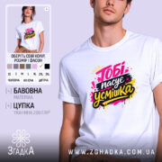 Футболка тобі пасує усмішка. Біла футболка на молодому чоловікові, зображена в анфас. Фон світло-фіолетовий, принт яскравий з написом 'Тобі пасує усмішка' в жовтому, чорному та рожевому кольорах, з кольоровими краплями.