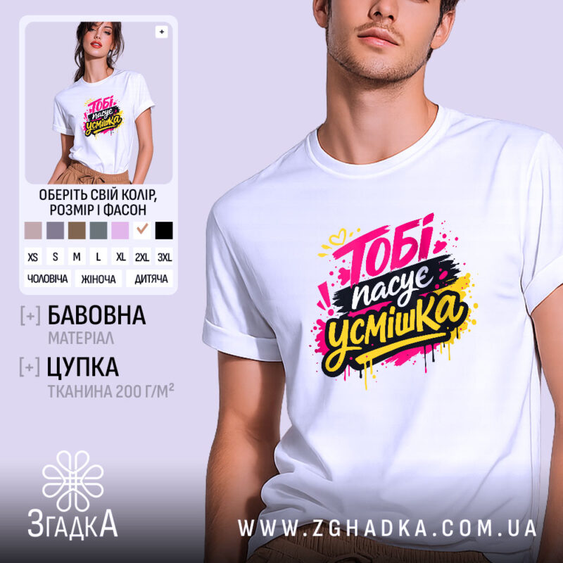 Футболка тобі пасує усмішка. Біла футболка на молодому чоловікові, зображена в анфас. Фон світло-фіолетовий, принт яскравий з написом 'Тобі пасує усмішка' в жовтому, чорному та рожевому кольорах, з кольоровими краплями.