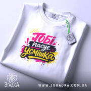 Футболка тобі пасує усмішка. Біла футболка, розкладена на поверхні, з принтом 'Тобі пасує усмішка' в яскравих кольорах. Фон світло-фіолетовий, видно етикетку з логотипом.