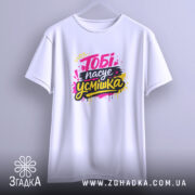 Футболка тобі пасує усмішка. Біла футболка, підвішена на вішалці, з принтом 'Тобі пасує усмішка' в яскравих кольорах. Фон світло-фіолетовий, футболка має класичний крій.