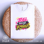 Футболка тобі пасує усмішка. Біла футболка, складена на плетеній кошику, з принтом 'Тобі пасує усмішка' в яскравих кольорах. Фон світлий, створює затишну атмосферу.