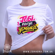 Футболка тобі пасує усмішка. Біла футболка, розкладена на поверхні, з принтом 'Тобі пасує усмішка' в яскравих кольорах. Фон світло-фіолетовий, видно етикетку з логотипом.