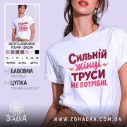 Футболка труси не потрібні. Біла футболка з принтом на грудях, на якому написано 'Сильний жінці труси не потрібні' червоним і рожевим кольорами, з жовтими акцентами. Модель позує з легким усмішкою, фон світло-сірий.
